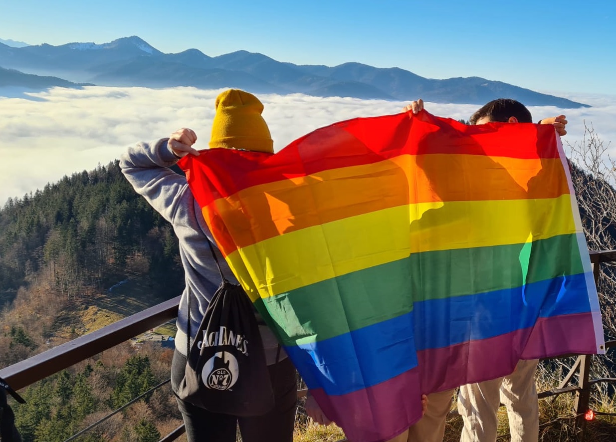 Zwei Teilnehmende stehen mit dem Rücken zur Kamera auf einem Berggipfel und halten eine Pride-Flagge zusammen hinter sich. Sie blicken auf eine Bergkette und einen klaren blauen Himmel.